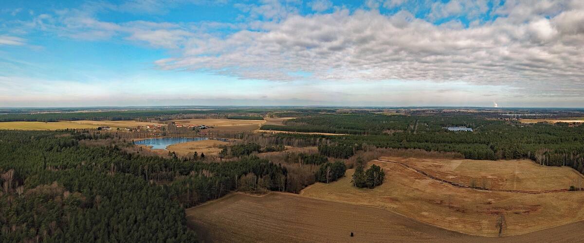 A rural landscape with a lake, dense forests, open fields, and distant buildings.