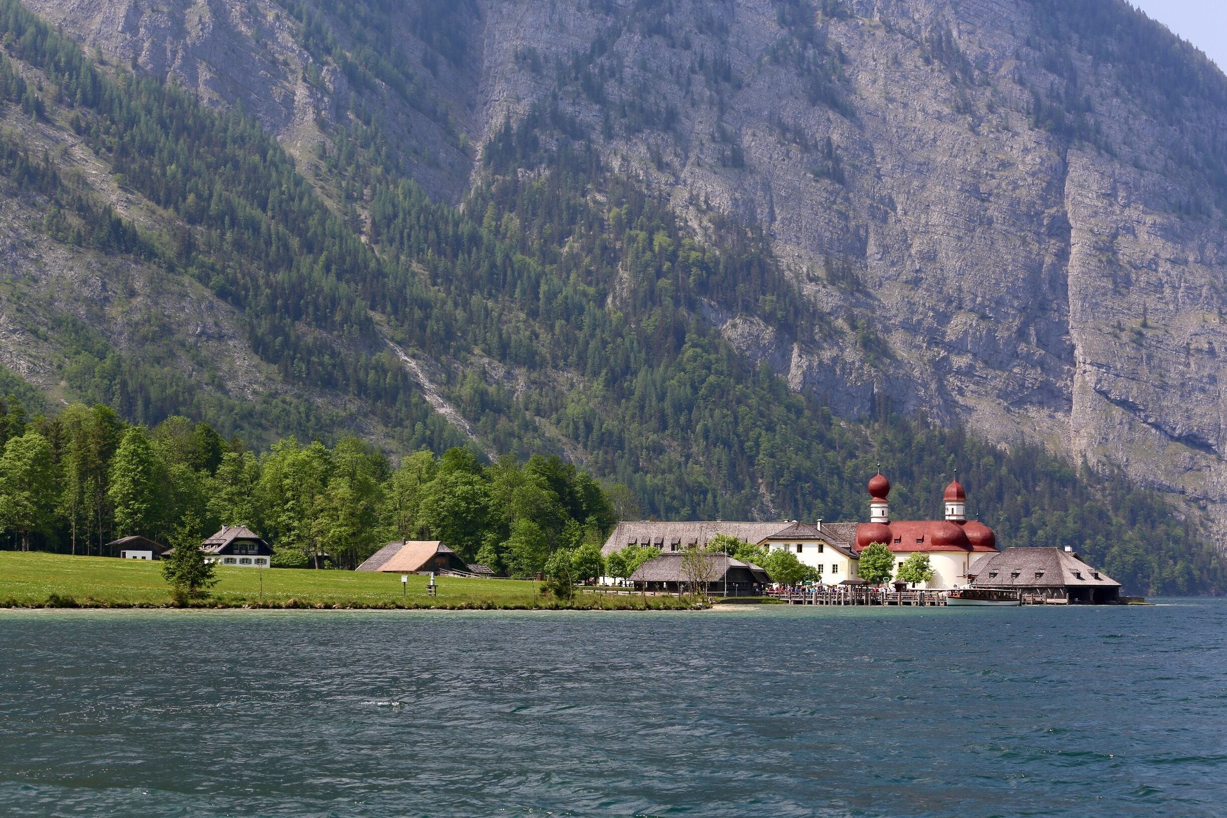 A lakeside building with red domes, surrounded by a forest and mountains.