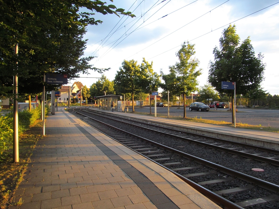 A train station platform with tracks, overhead wires, and a pedestrian walkway.