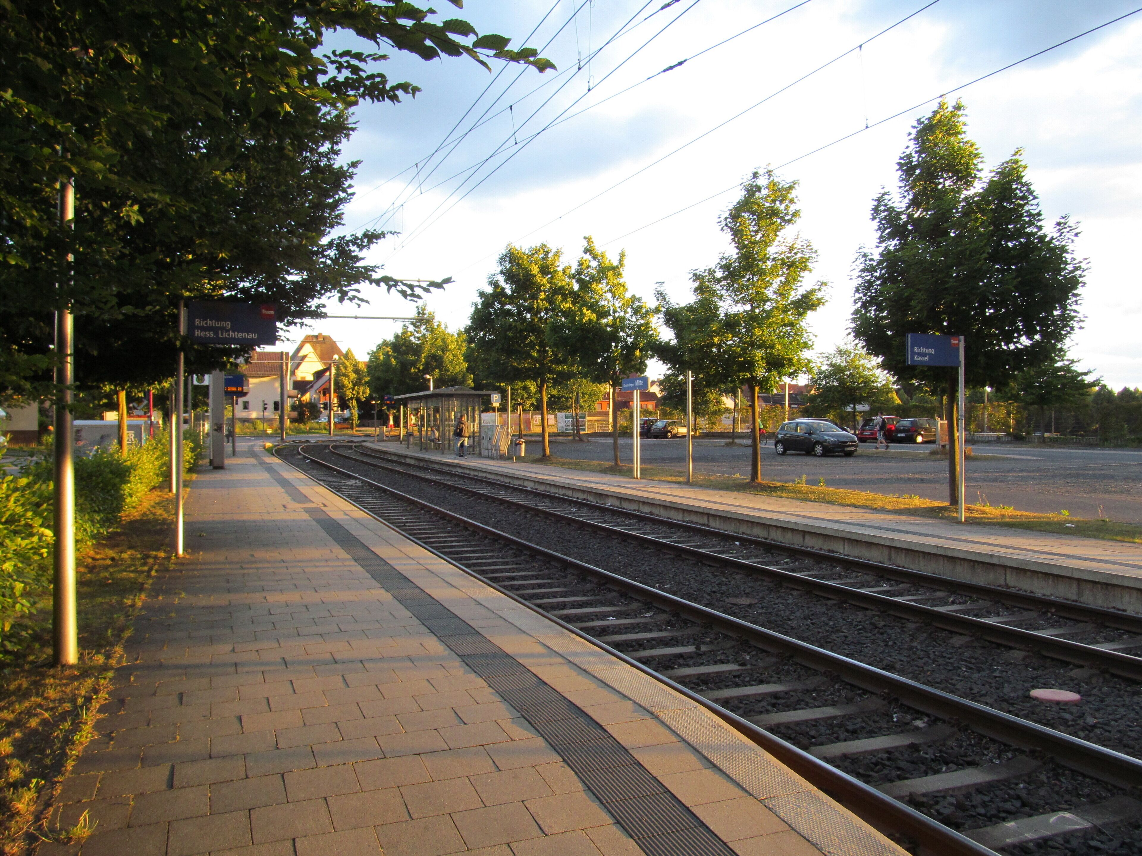 A train station platform with tracks, overhead wires, and a pedestrian walkway.