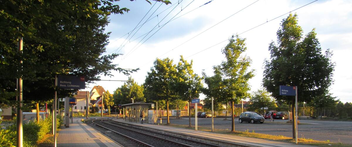 A train station platform with tracks, overhead wires, and a pedestrian walkway.