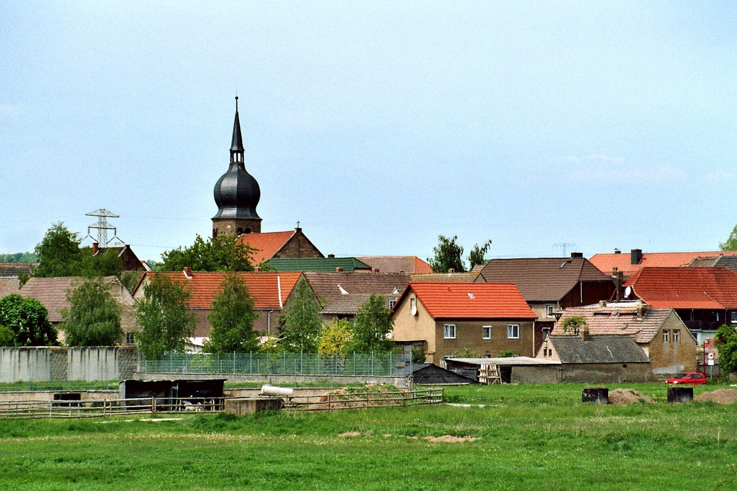 A rural village with a prominent church steeple, houses with red roofs, and a fenced area with a small building.