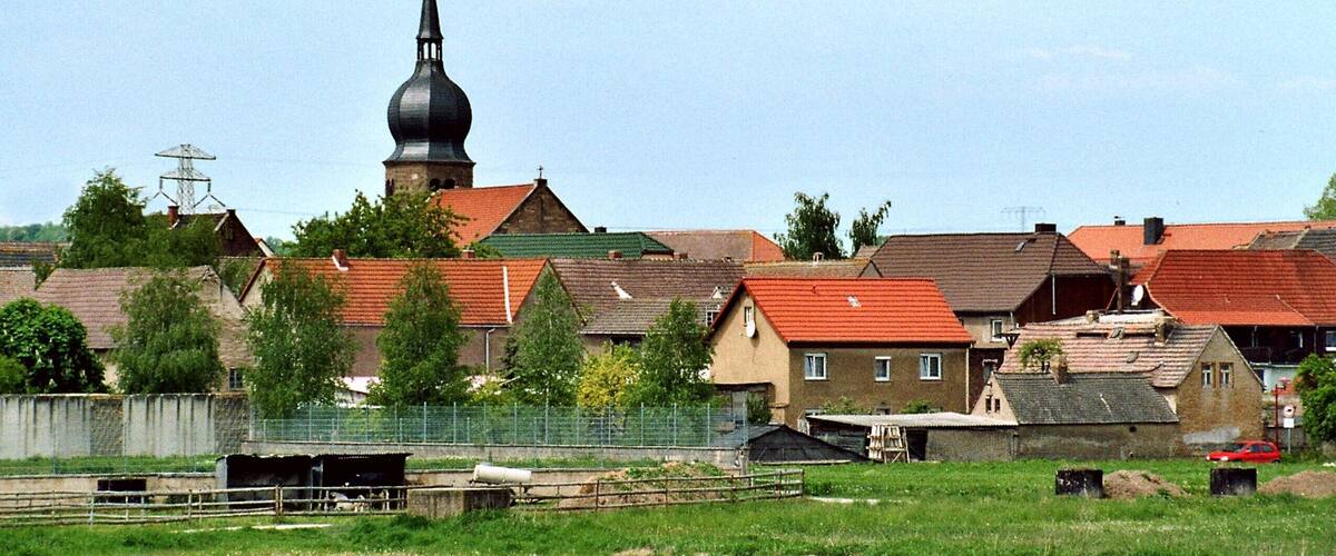 A rural village with a prominent church steeple, houses with red roofs, and a fenced area with a small building.