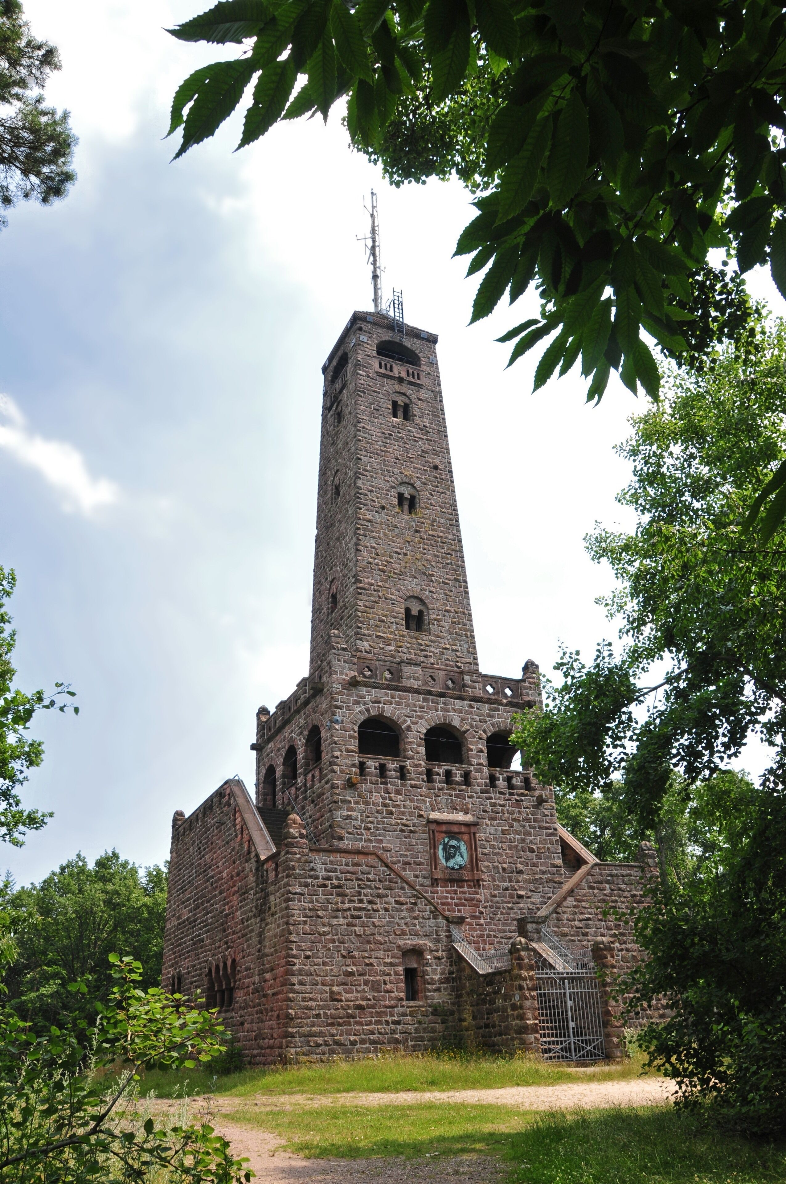 A tall stone tower with a clock and antenna, surrounded by trees.