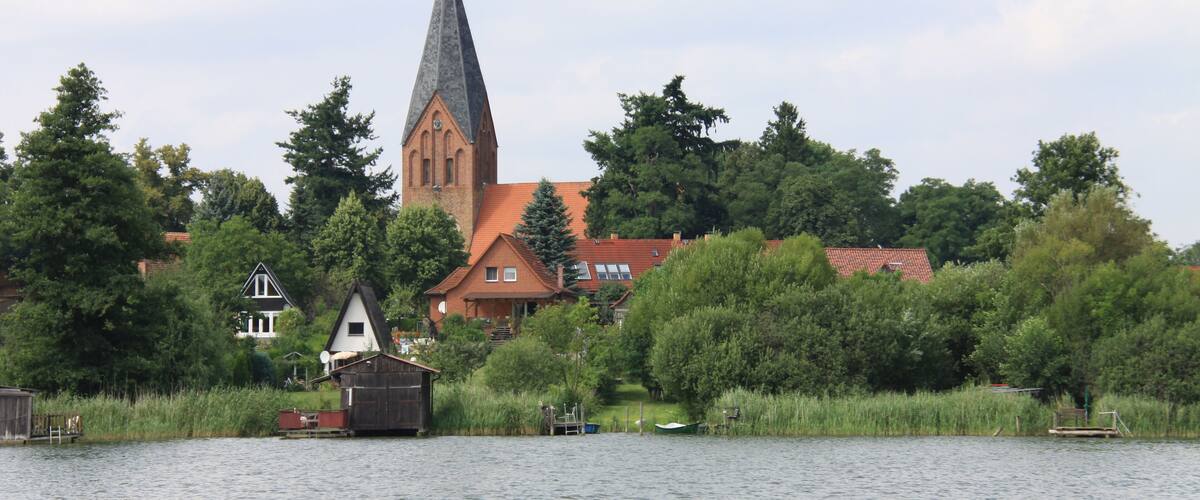 A church with a tall spire, surrounded by trees and houses by a lake.