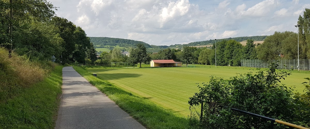 A paved path, a green field, a small building, and trees under a partly cloudy sky.