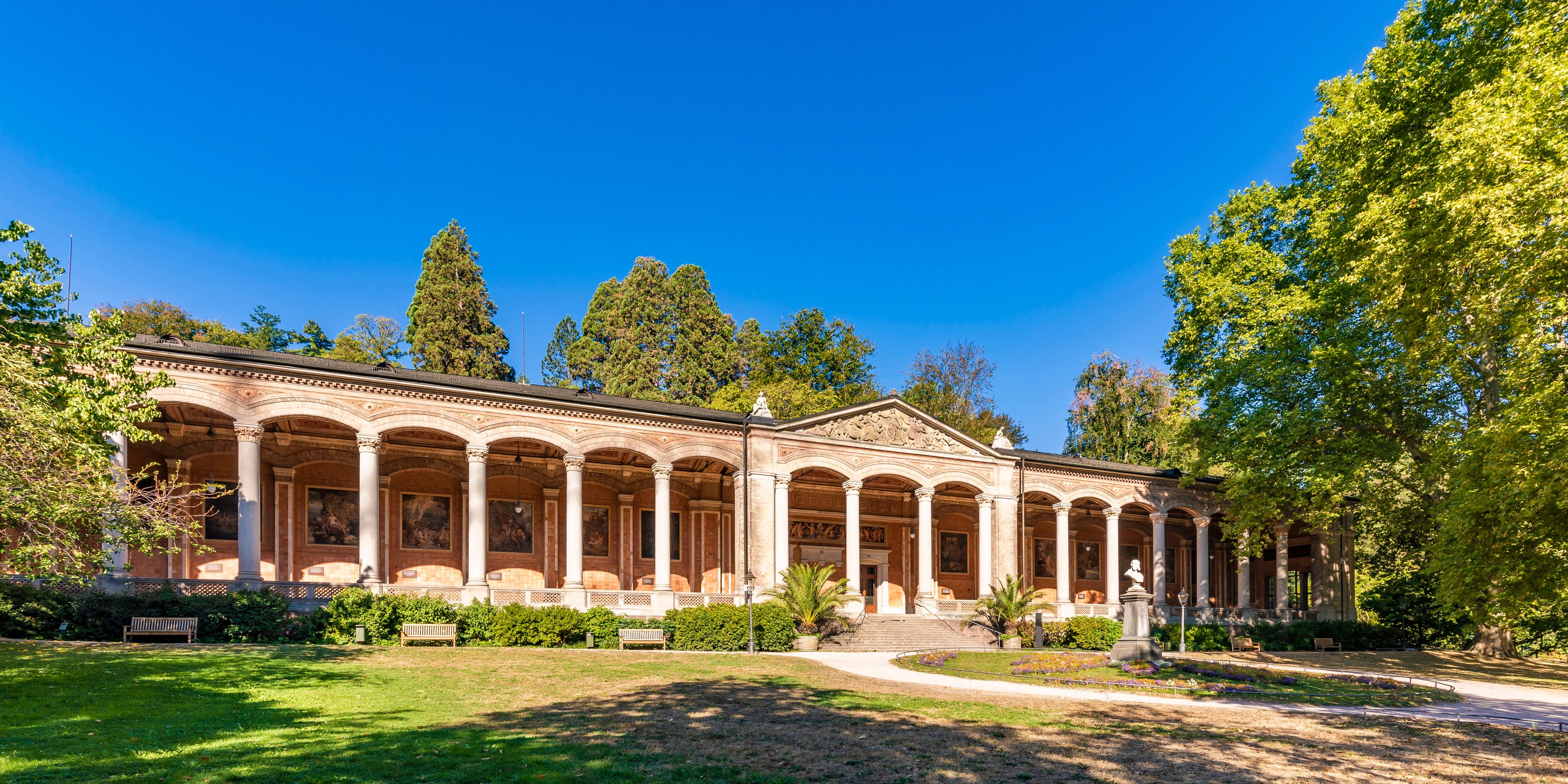 A classical building with a colonnade, surrounded by a well-maintained garden and trees.