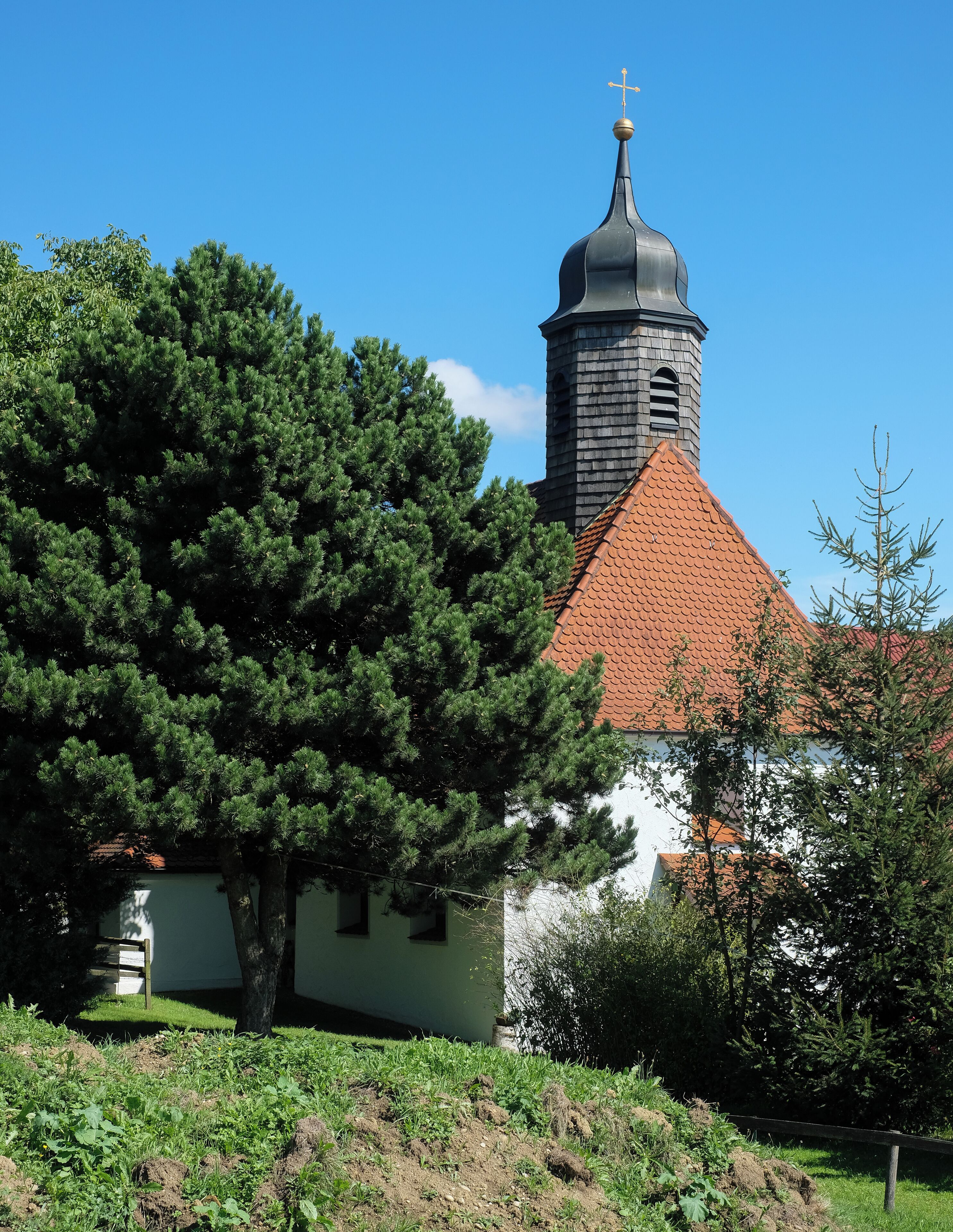 A church with a red roof and a tall tower with a cross on top, surrounded by trees and greenery.