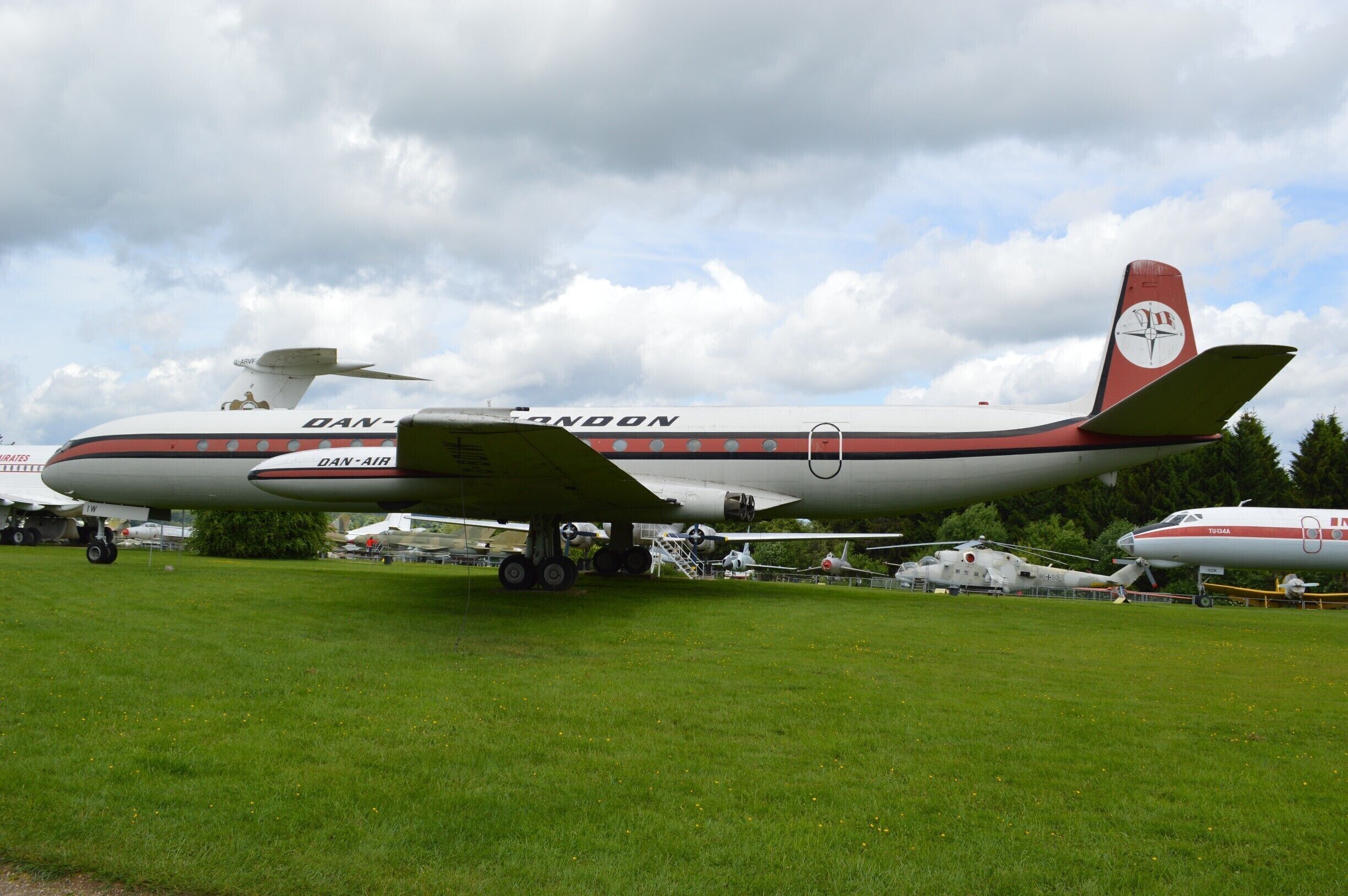 A vintage airplane with the text "DAN-AIR" on its fuselage, parked on a grassy field with other aircraft in the background.