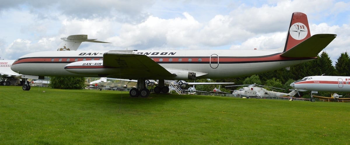 A vintage airplane with the text "DAN-AIR" on its fuselage, parked on a grassy field with other aircraft in the background.