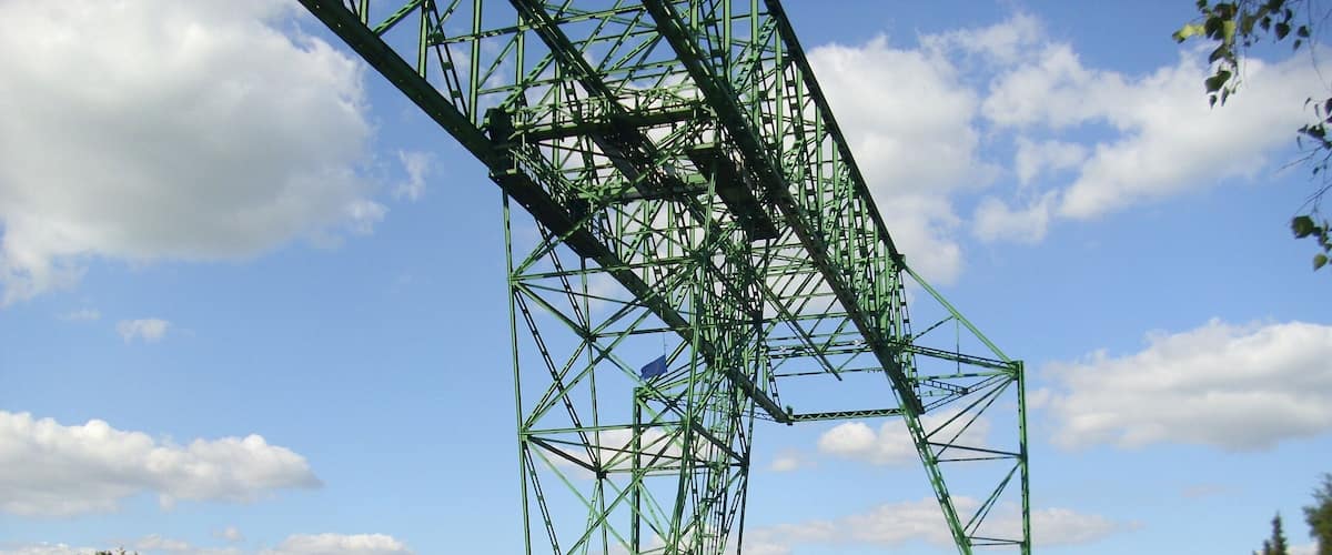A green metal bridge over a river with houses and trees in the background.