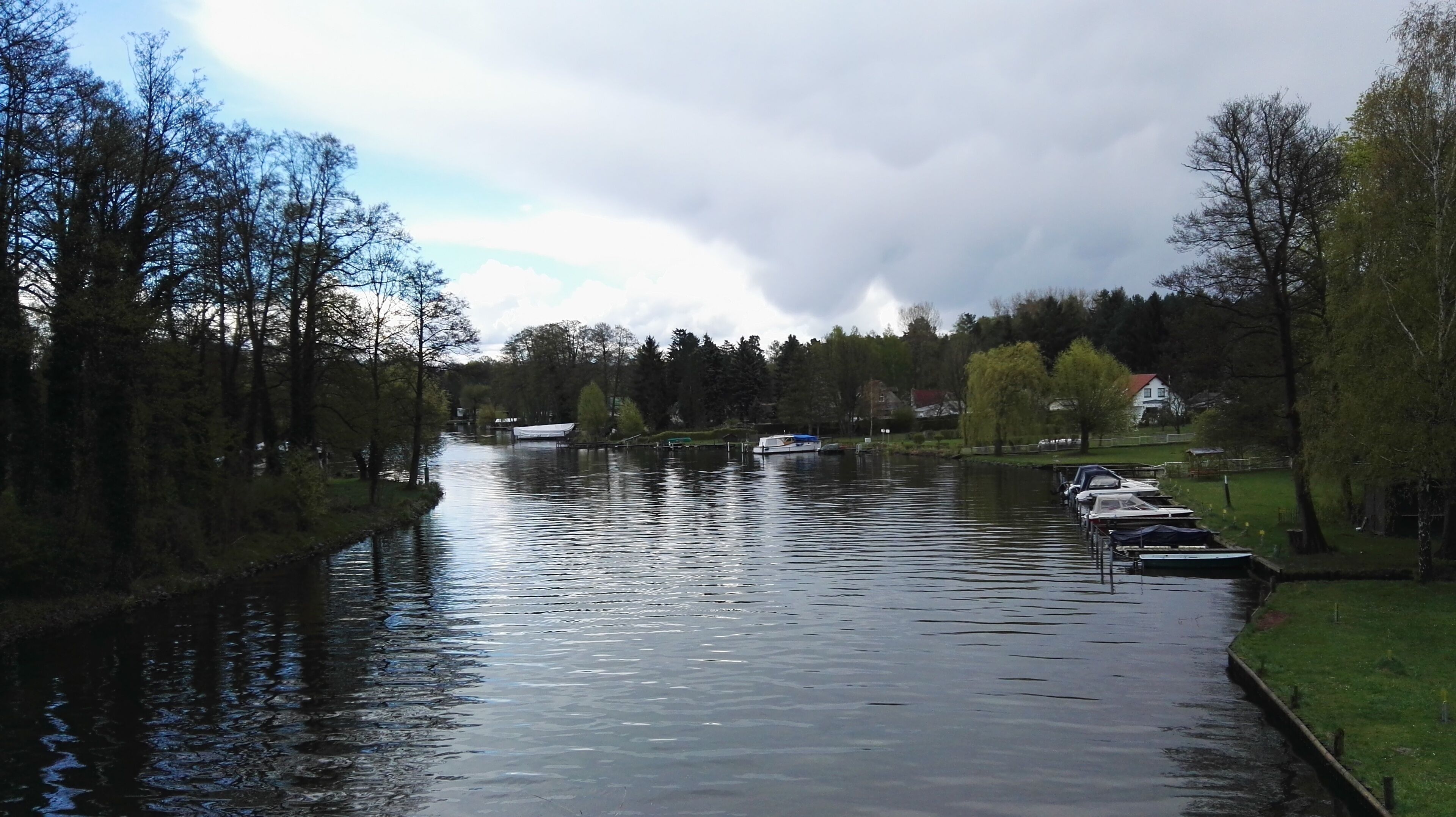 A calm river with boats docked on the right bank, surrounded by trees and a grassy area.