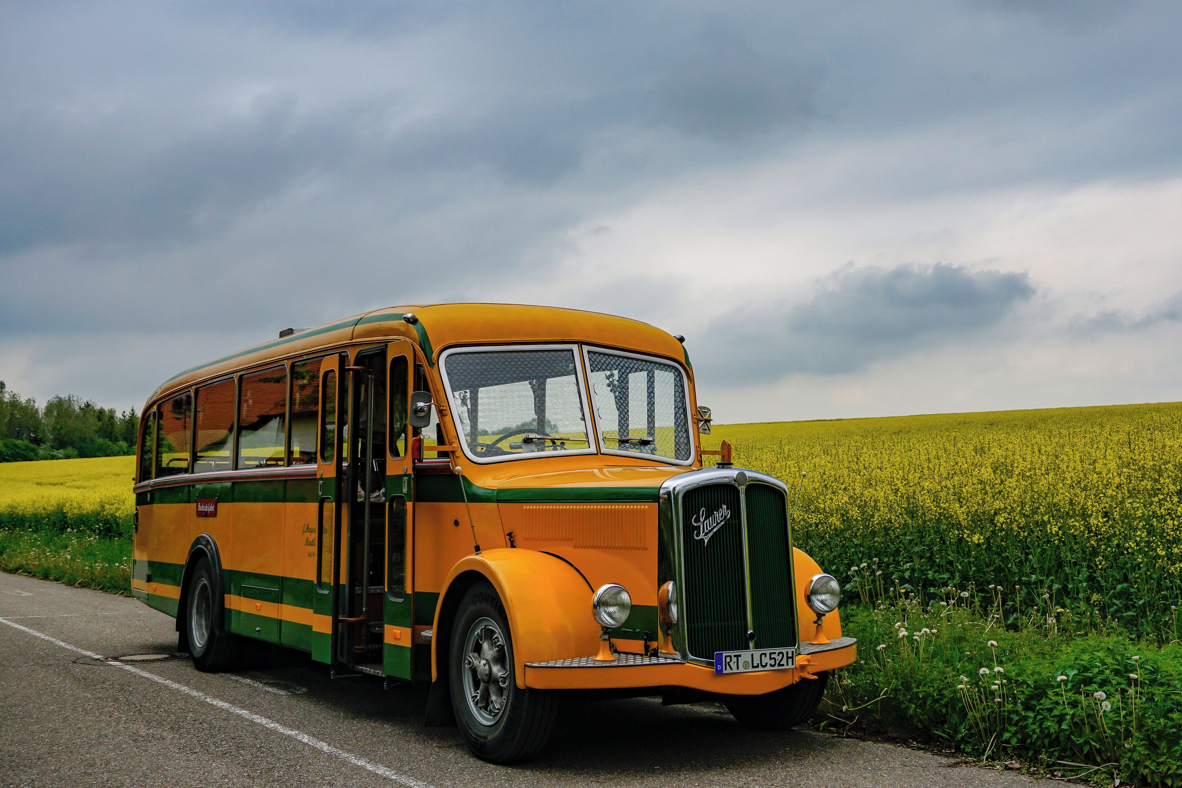 An orange vintage bus parked on a road with a field of yellow flowers in the background.