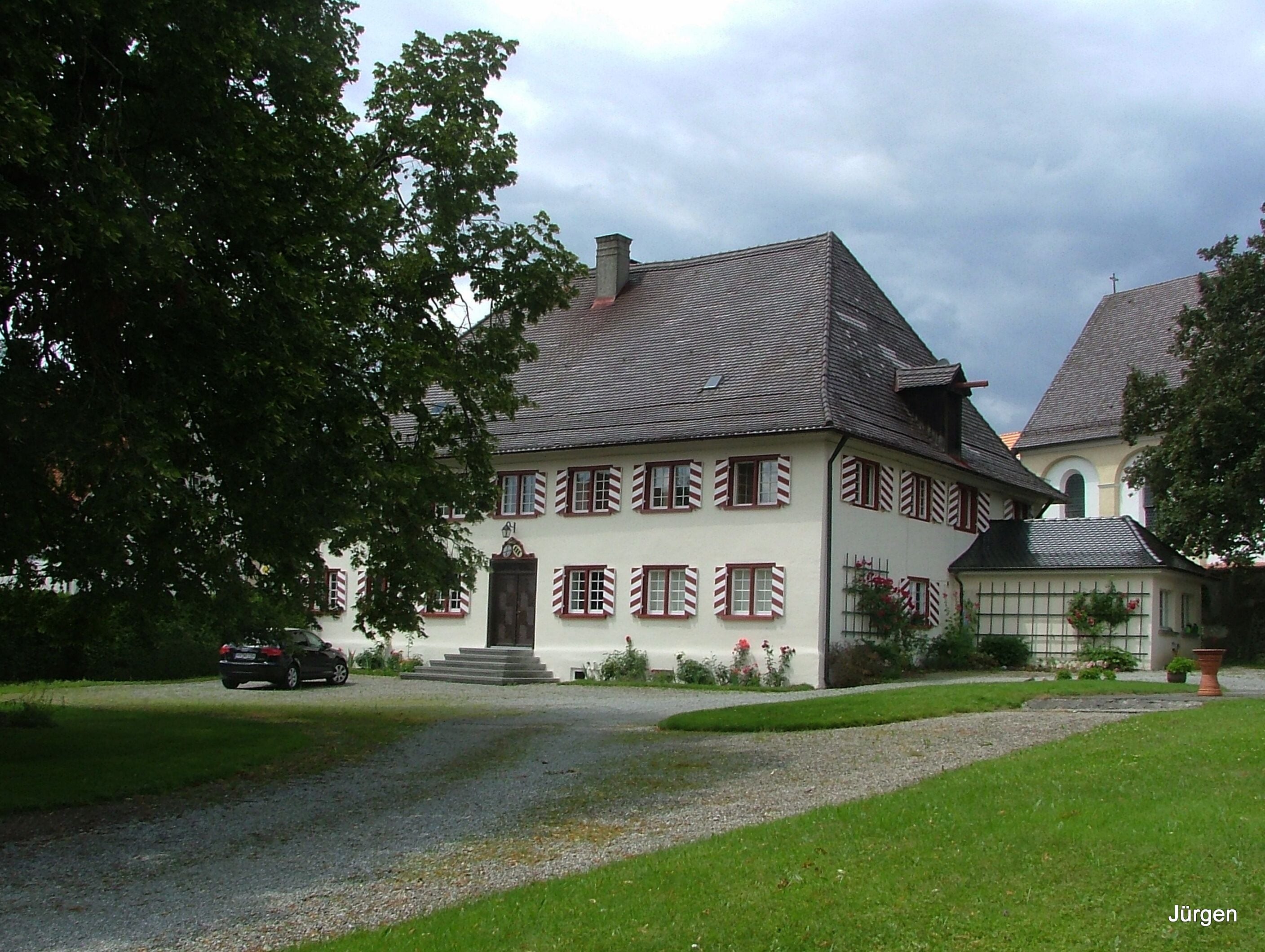 A large, traditional house with a steeply pitched roof, surrounded by a well-maintained lawn and a gravel driveway.