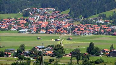 A village with red-roofed houses nestled in a valley surrounded by green fields and forested hills.