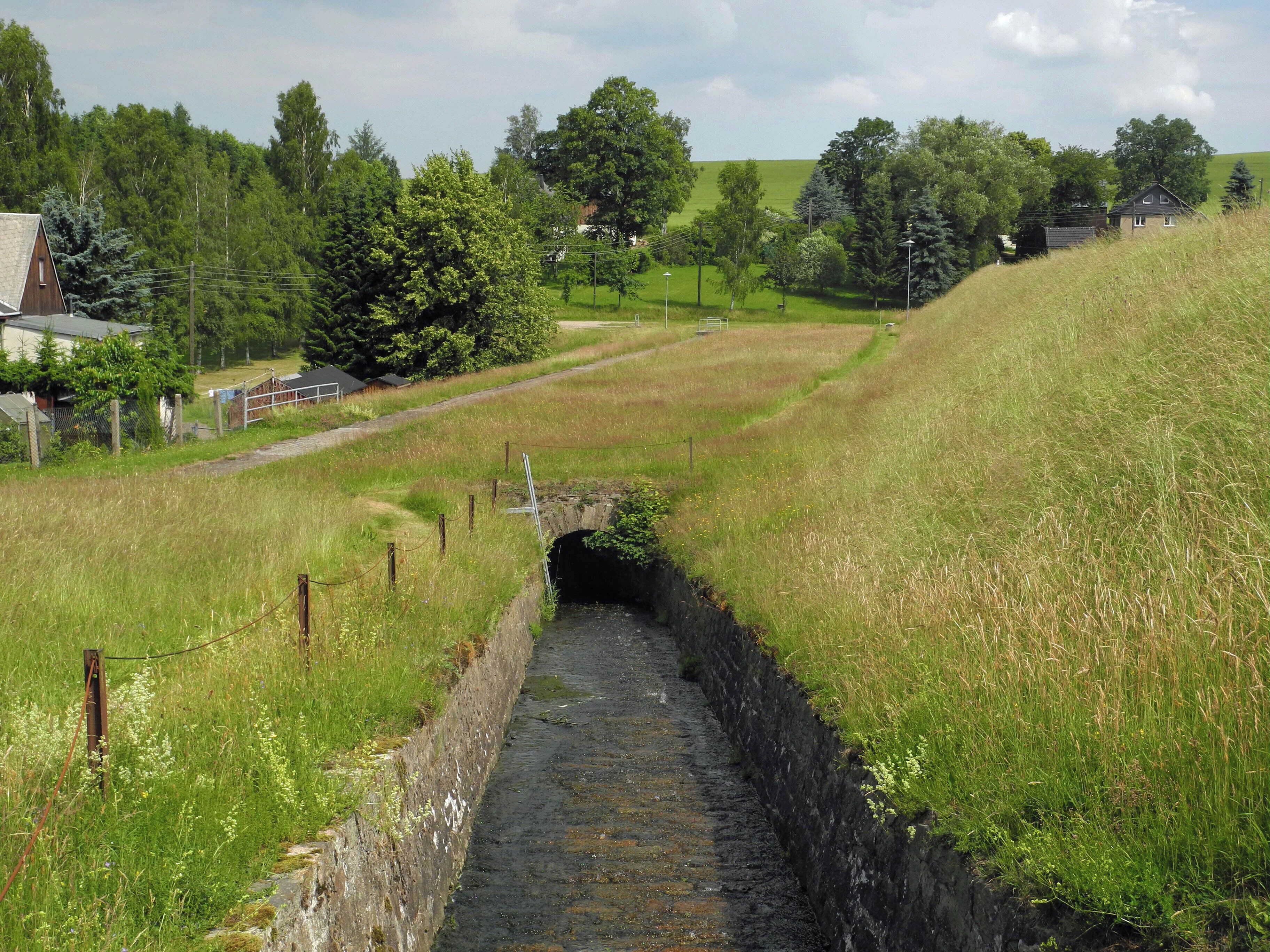 A rural landscape with a narrow stone water channel, grassy fields, and a few scattered trees.