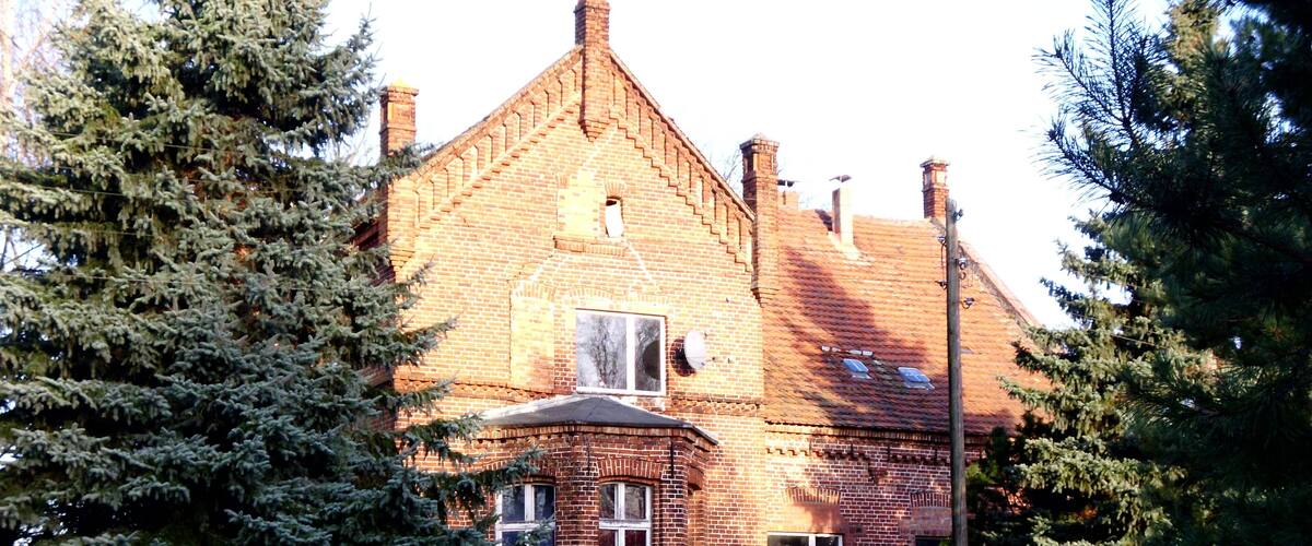 A large brick house with a steeply pitched roof, surrounded by evergreen trees and a grassy yard.