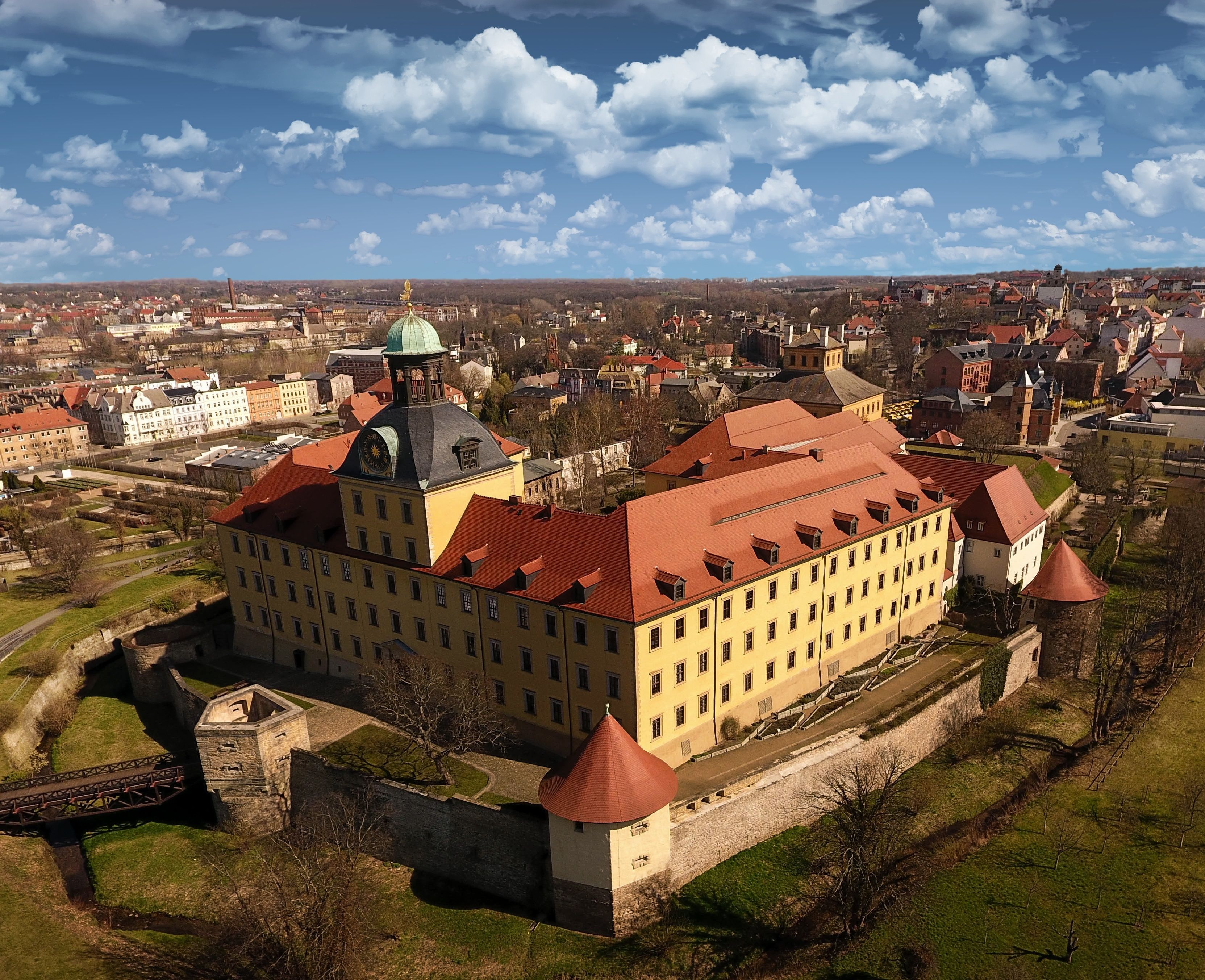 Ein historisches Gebäude mit rotem Dach und grüner Kuppel, umgeben von einer befestigten Mauer und einem Burggraben.