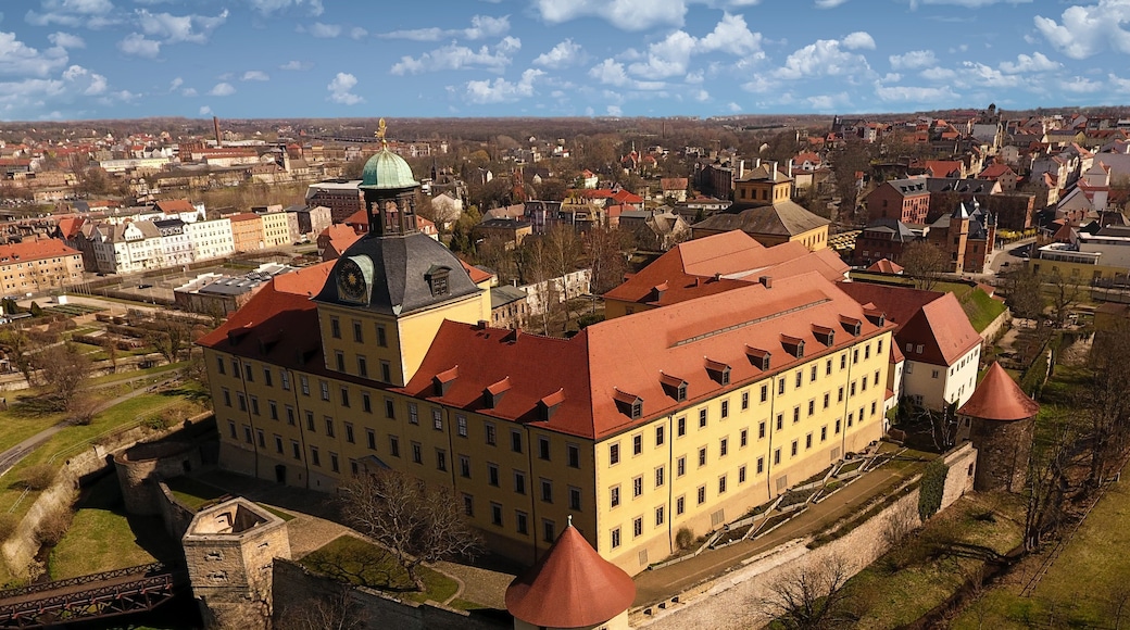 Ein historisches Gebäude mit rotem Dach und grüner Kuppel, umgeben von einer befestigten Mauer und einem Burggraben.