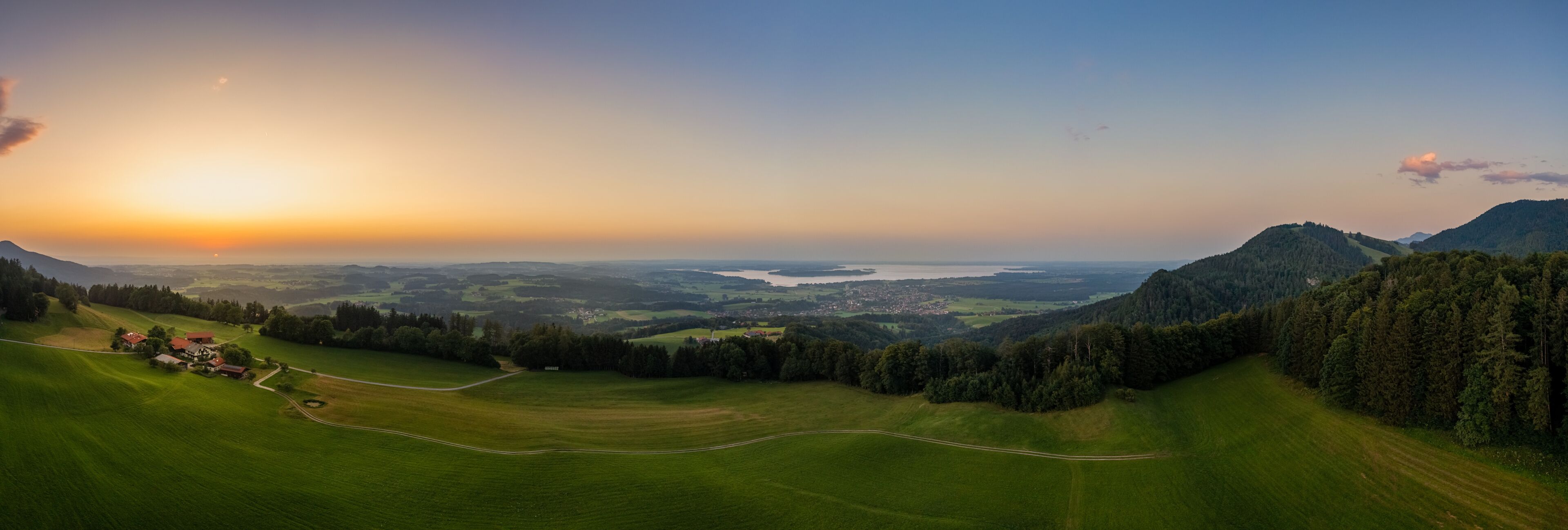 A panoramic view of a lush green valley with a winding road, a small settlement, and a distant body of water under a sunset sky.