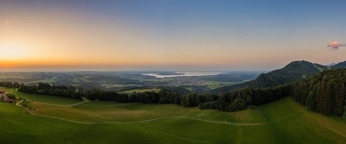 A panoramic view of a lush green valley with a winding road, a small settlement, and a distant body of water under a sunset sky.
