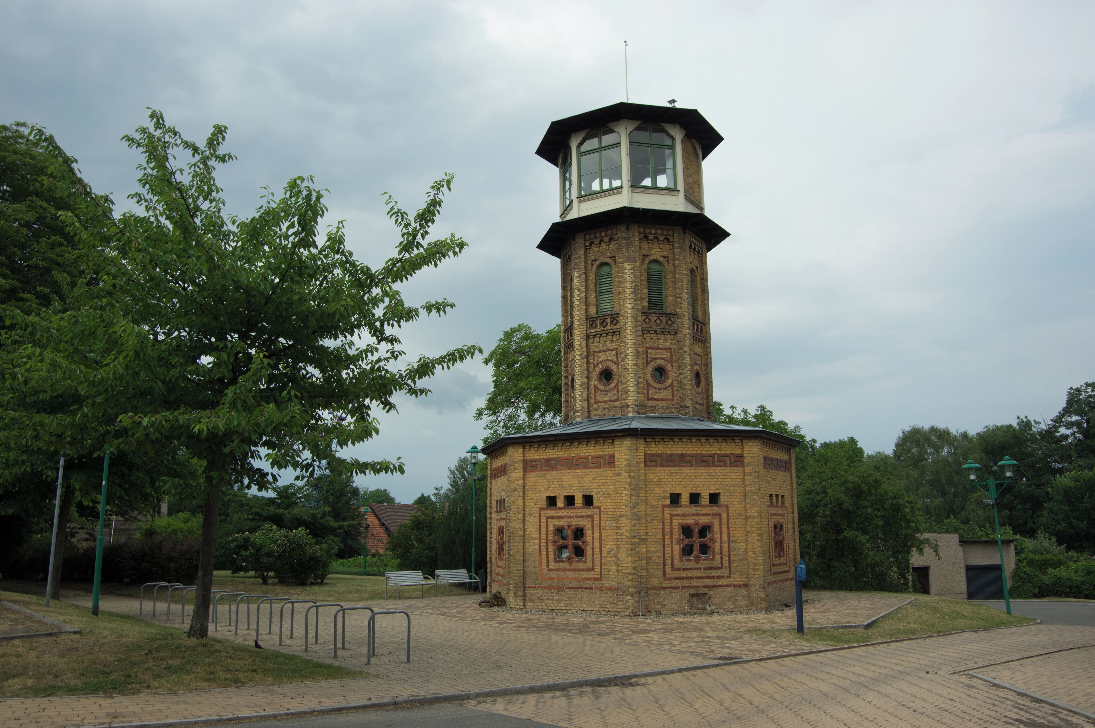 A brick tower with a clock and a small balcony, surrounded by trees and benches.