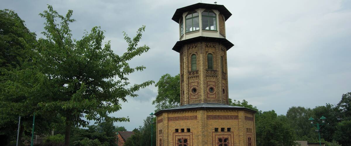 A brick tower with a clock and a small balcony, surrounded by trees and benches.