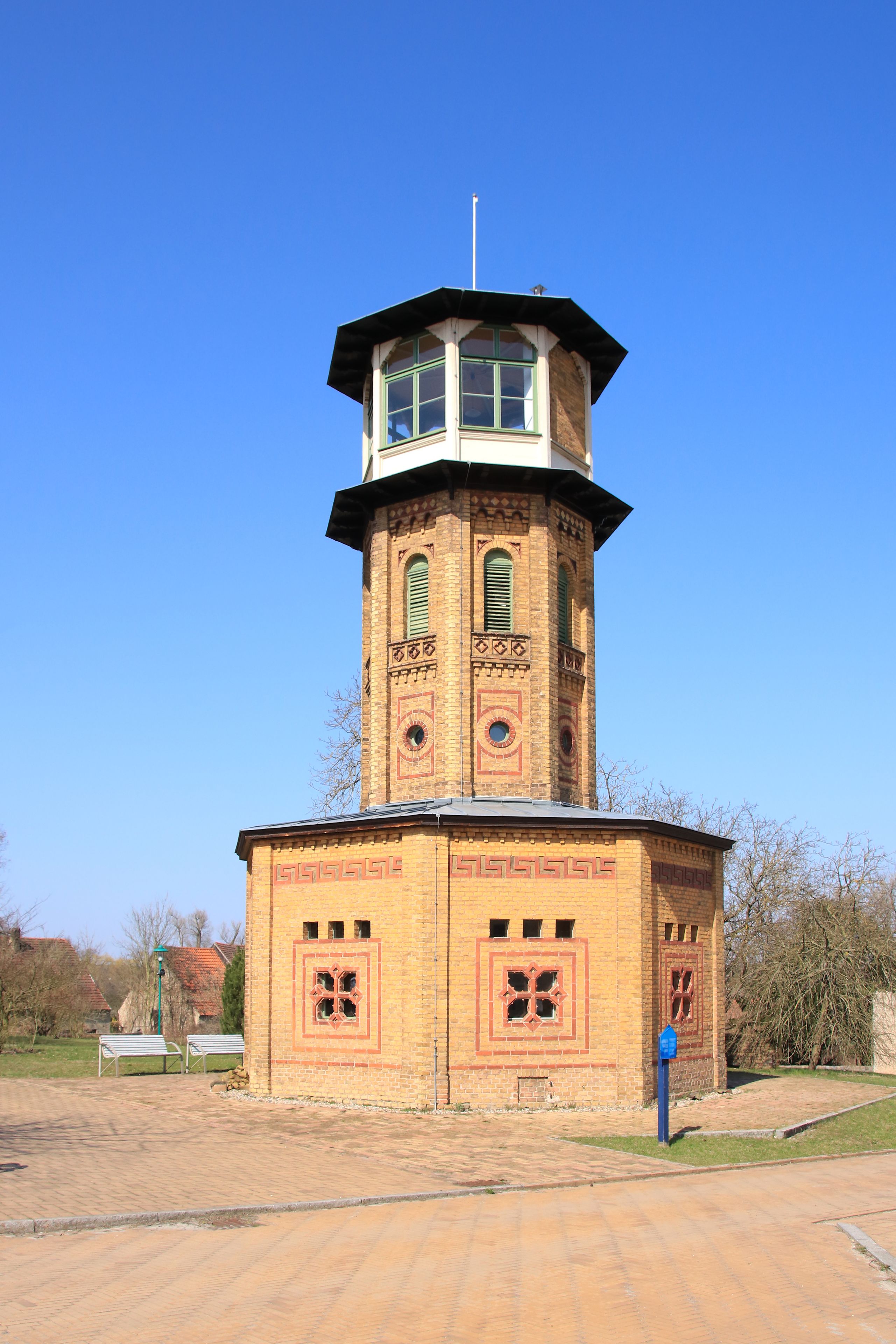 A brick tower with a clock and a flagpole on top, surrounded by a paved area and trees.