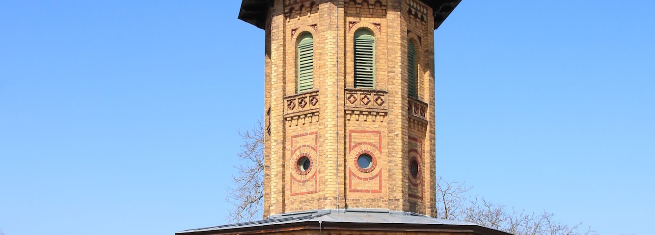 Uma torre de tijolos com um relógio e um mastro, rodeada por um espaço pavimentado e árvores.