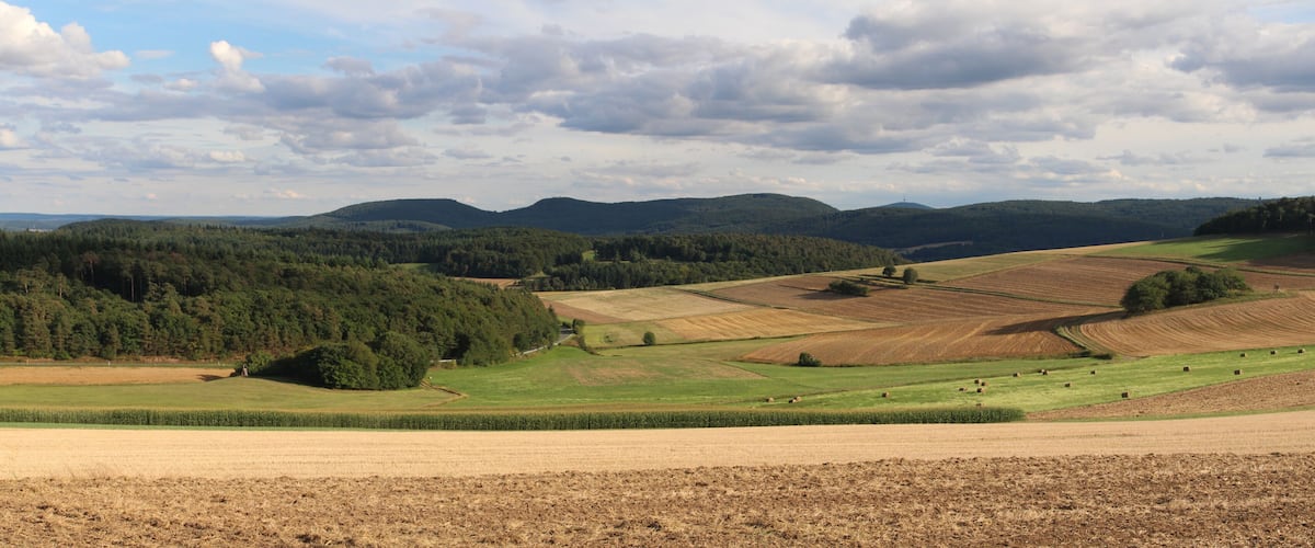 A rural landscape with fields, forests, and rolling hills under a partly cloudy sky.