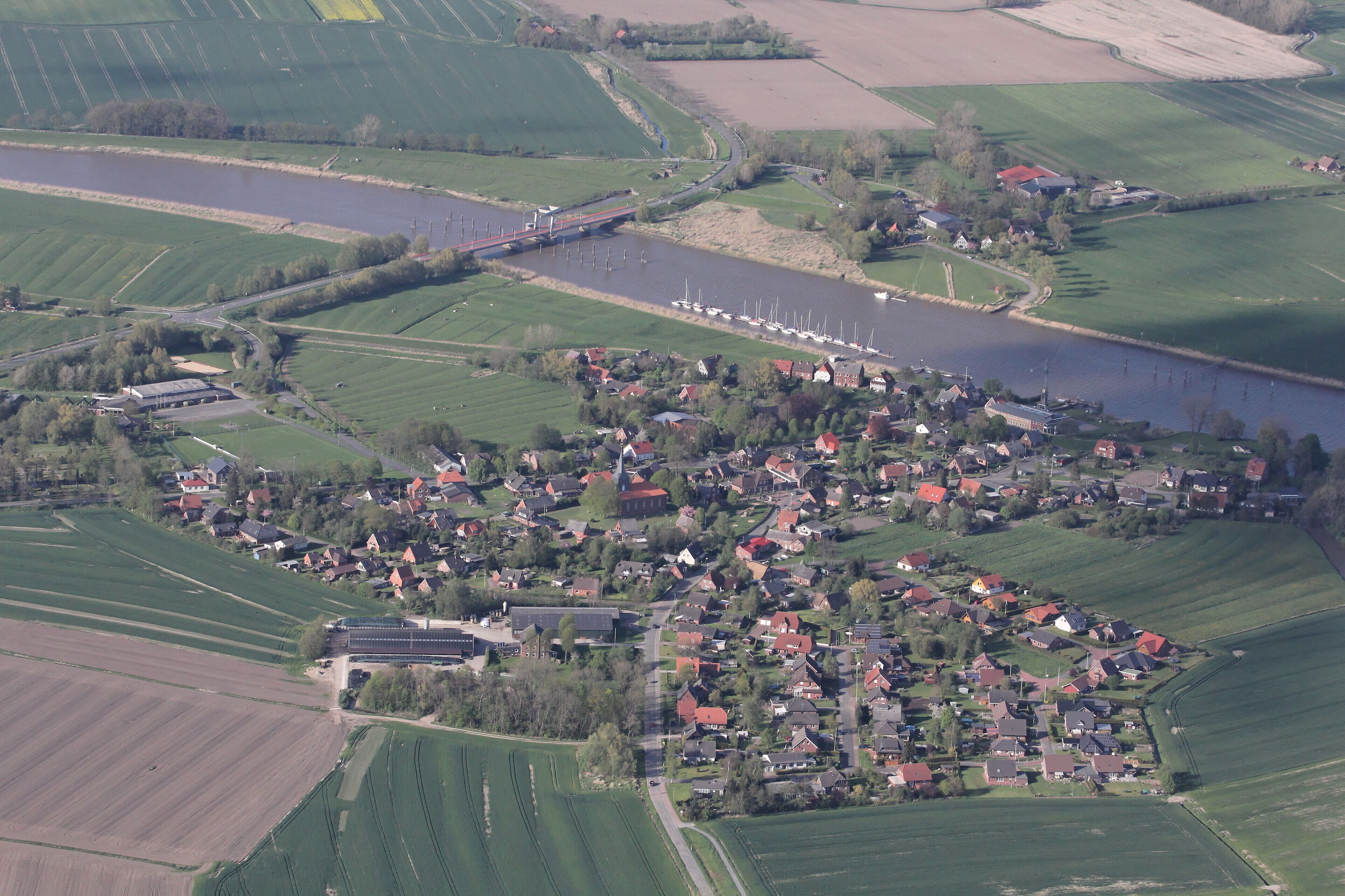 An aerial view of a small village with a river, fields, and a bridge.