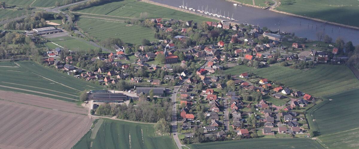 An aerial view of a small village with a river, fields, and a bridge.