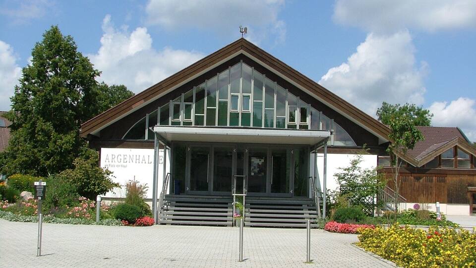 A modern building with a glass facade and a wooden roof, surrounded by a garden with flowers and trees.