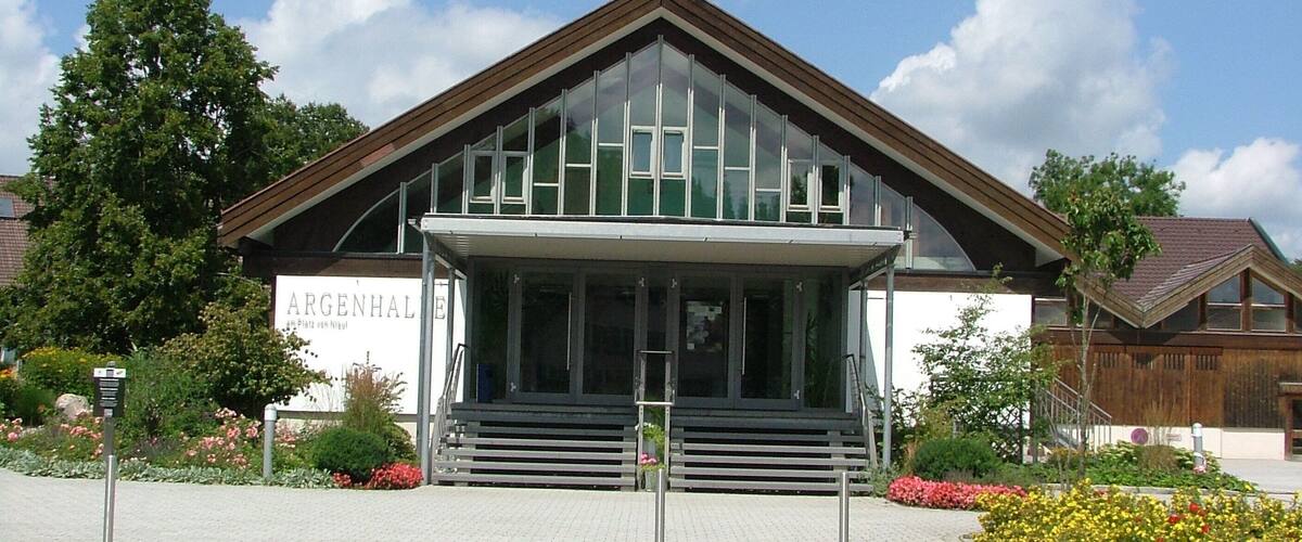 A modern building with a glass facade and a wooden roof, surrounded by a garden with flowers and trees.