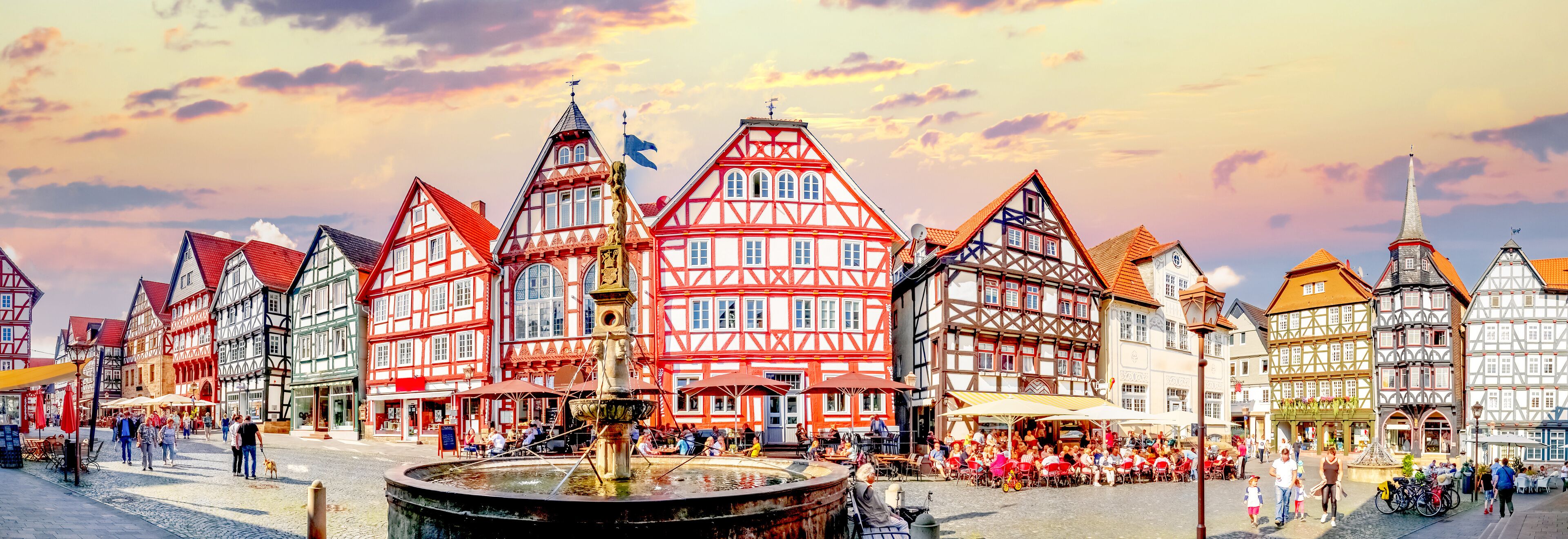 A European town square with traditional half-timbered buildings, a central fountain, and people gathered around outdoor seating areas.