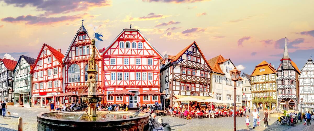 A European town square with traditional half-timbered buildings, a central fountain, and people gathered around outdoor seating areas.