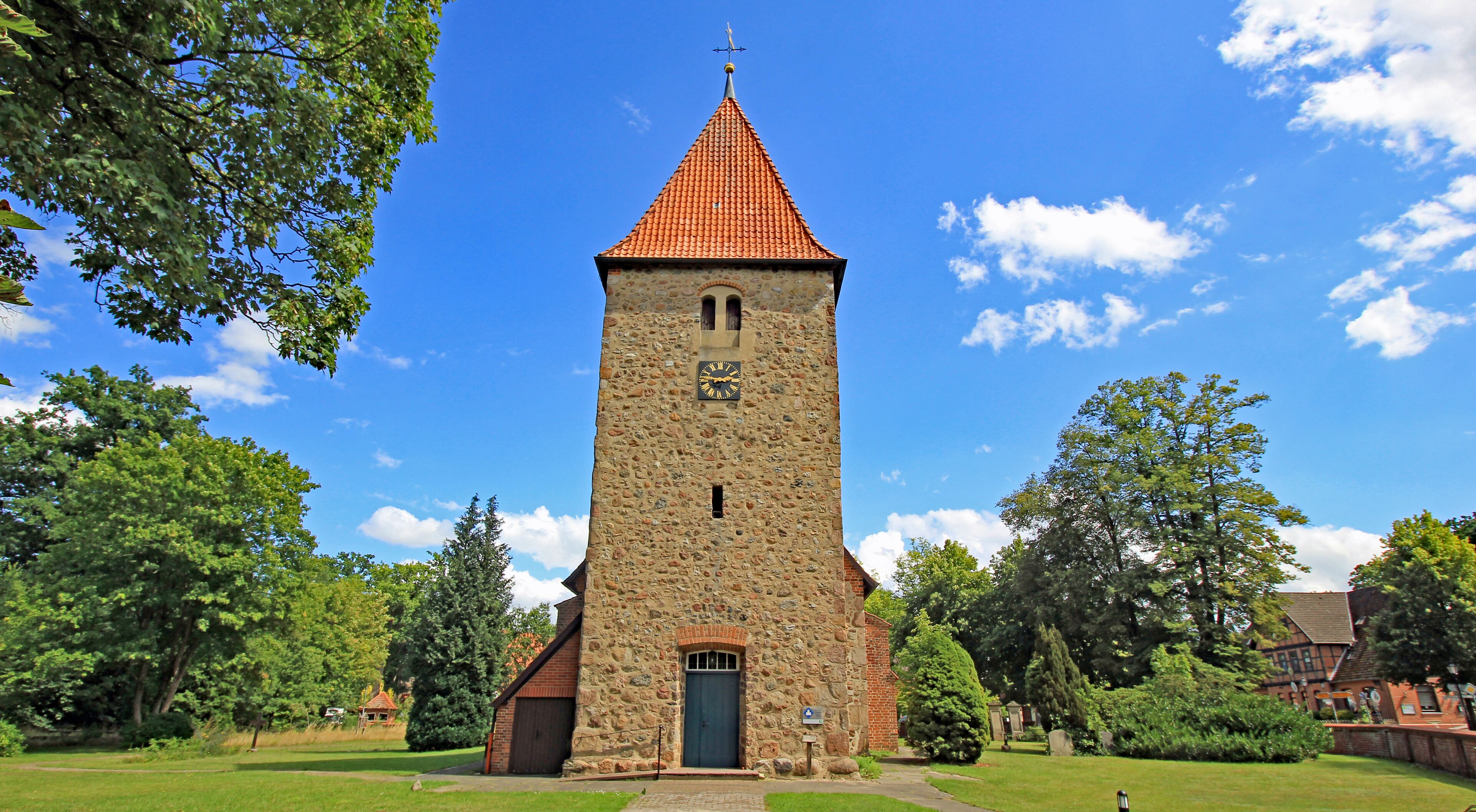 A church with a stone tower and a red roof, surrounded by trees and a clear sky.