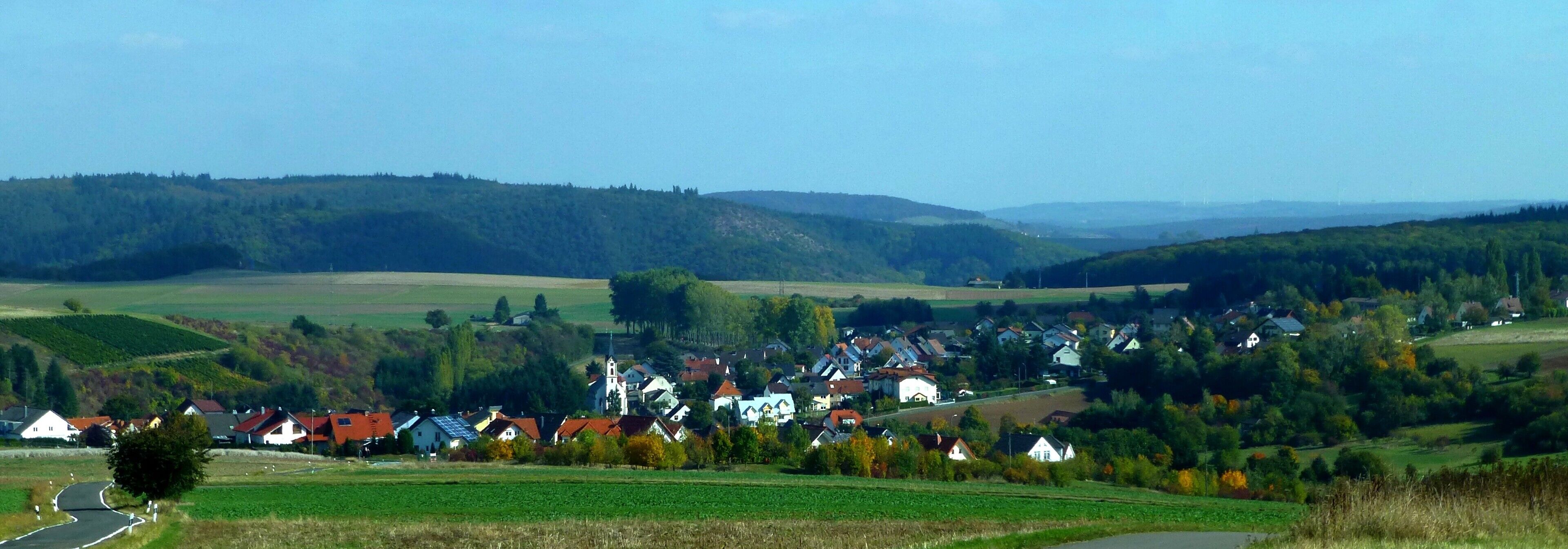 A rural village nestled in a valley with rolling hills and vineyards in the background.