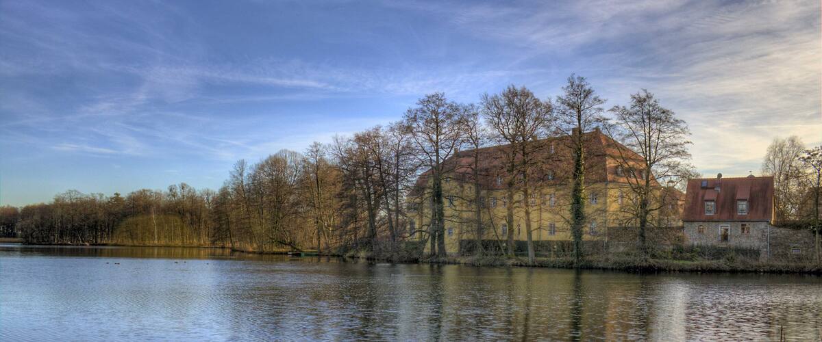 A calm lake with reflections of buildings and trees, surrounded by a clear sky.