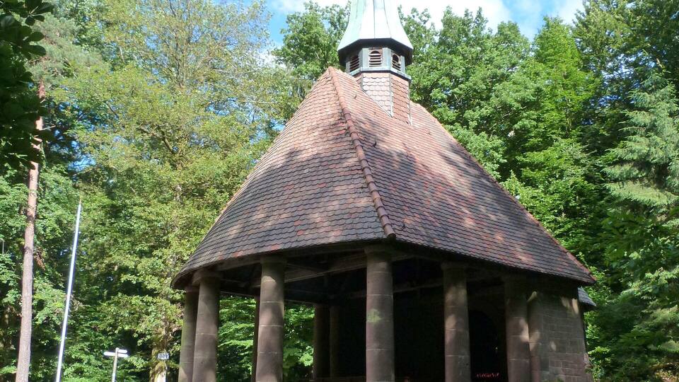 A small stone chapel with a tiled roof and a steeple, surrounded by lush greenery.