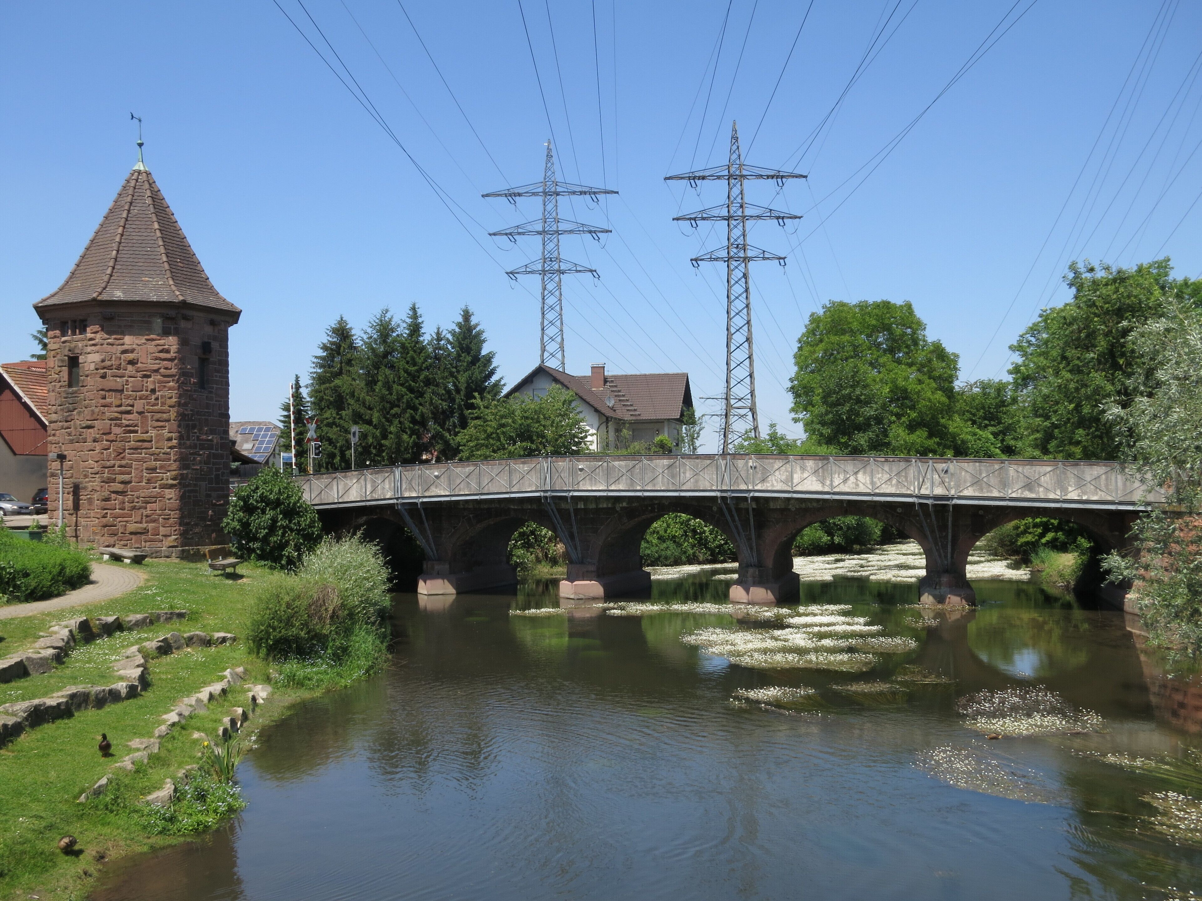 A stone bridge over a calm river, surrounded by greenery and a stone tower.