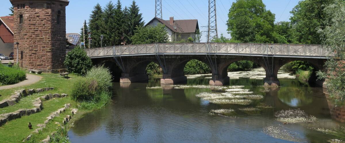 A stone bridge over a calm river, surrounded by greenery and a stone tower.