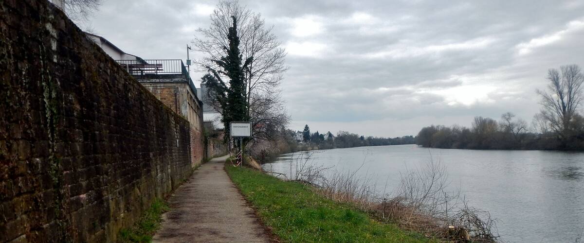 A riverside path with a brick wall on one side and a sign that reads "HAFENGEBET".