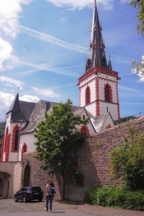 A church with a tall spire and red accents, a stone wall, and a tree in front.