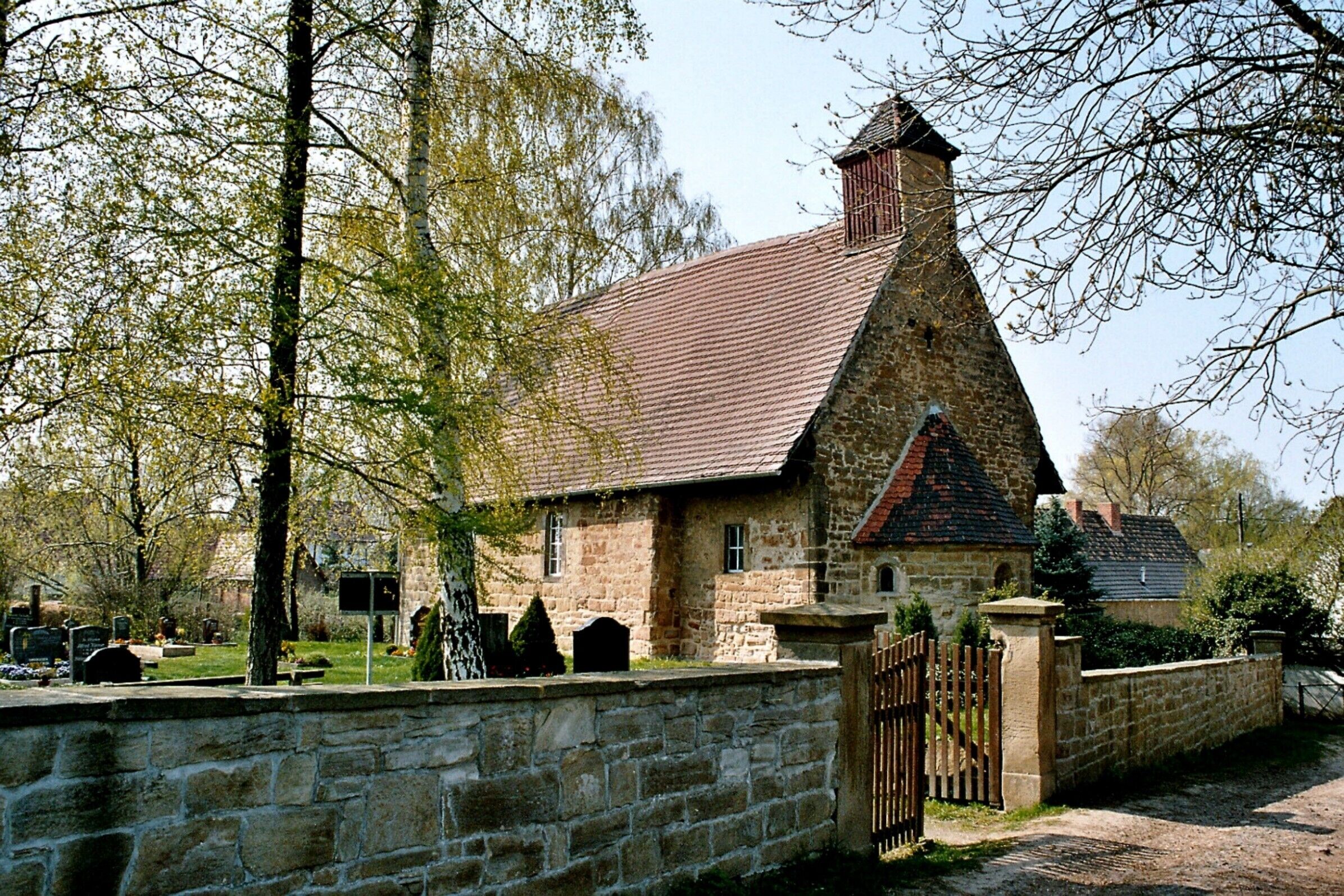 A stone church with a red tiled roof, surrounded by a cemetery and trees.