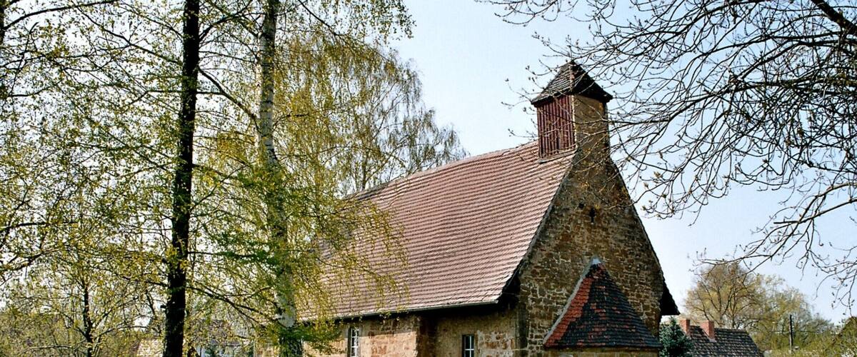 A stone church with a red tiled roof, surrounded by a cemetery and trees.