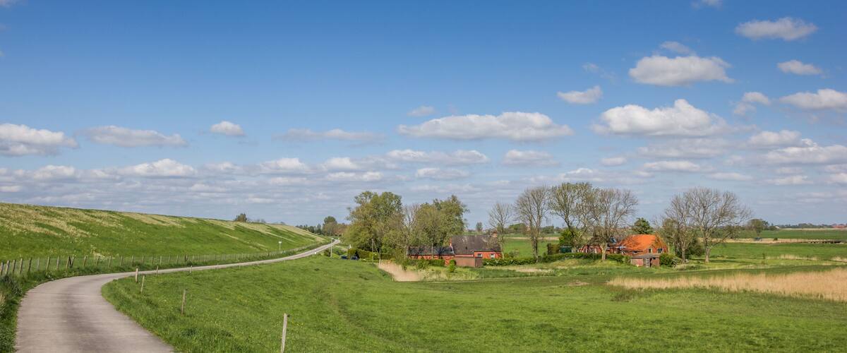 A rural landscape with a curving road, green fields, a few houses, and a clear blue sky with clouds.