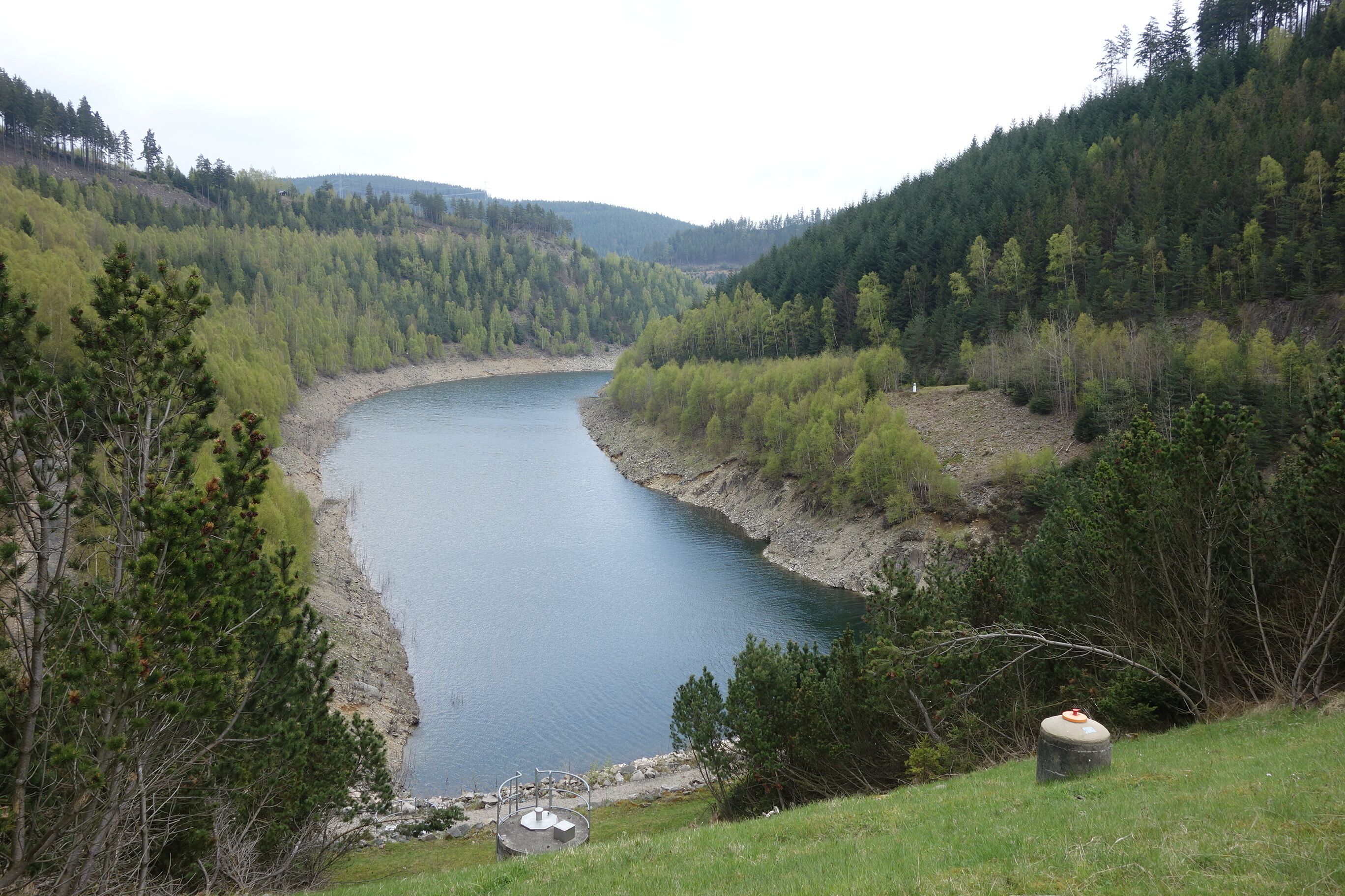 A river winding through a forested valley with a small dock and a trash can nearby.