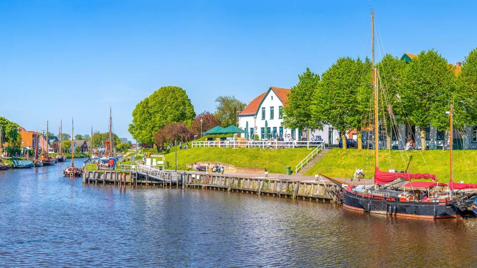 A canal with boats docked, a white building with a red roof, and a grassy embankment with trees.