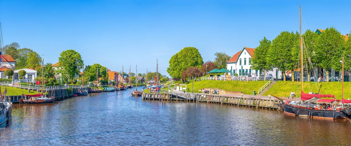 A canal with boats docked, a white building with a red roof, and a grassy embankment with trees.